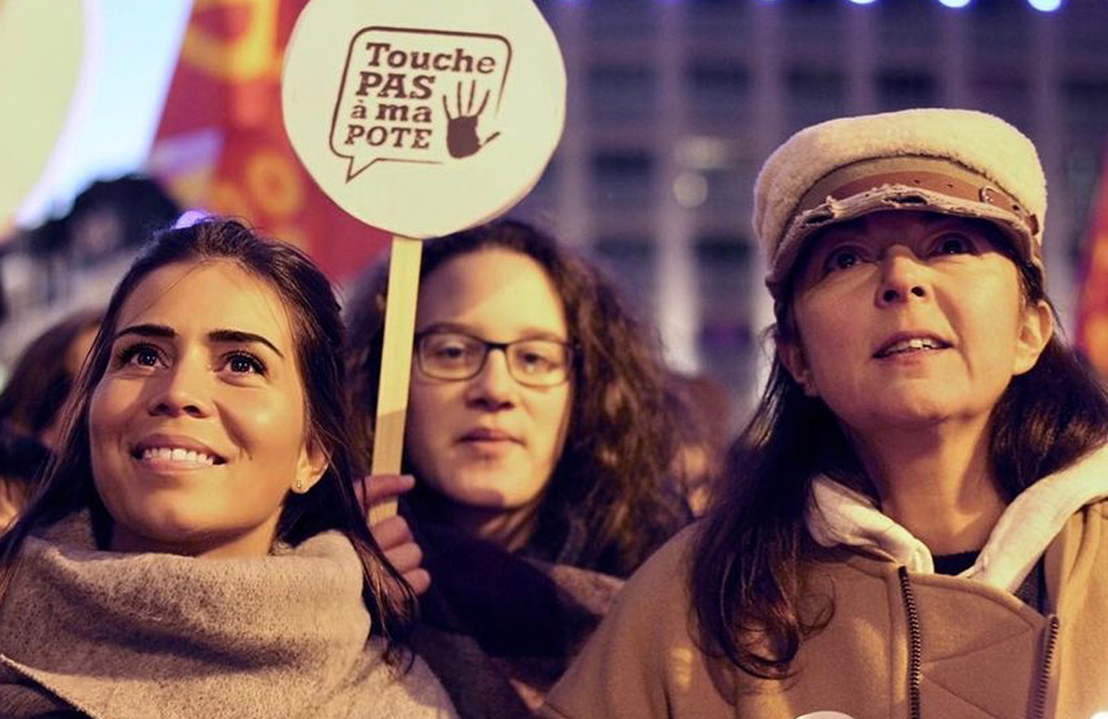 Trois femmes lors d'un rassemblement nocturne, l'une tenant une pancarte « Touche pas à ma pote » ornée d'une main